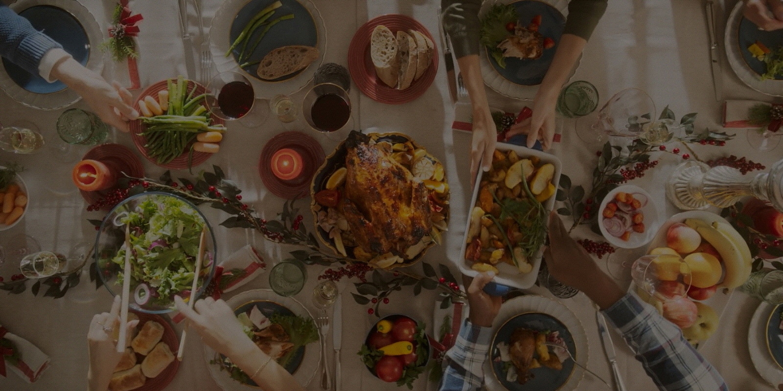 A dining table filled with various holiday dishes, fruits, and drinks, viewed from above, multiple hands are serving food.	