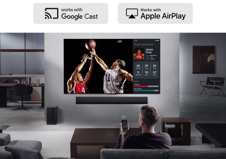 Person in their living room holding their phone. On the phone is a transmitting icon showing that the phone screen is being mirrored on the TV. On the TV is a basketball game to the side is the mirrored screen, showing player statistics.