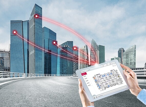 A person holds a tablet displaying a control solution. Four red lines extend from the tablet, connecting to buildings on the left.