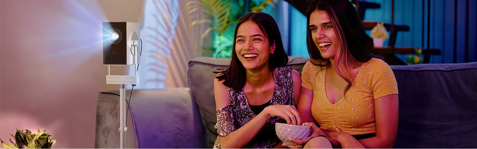 Two women are sitting on a sofa, eating popcorn and laughing while watching content. Next to them, a Cinebeam Q is placed on a stand, projecting the video onto a screen.	