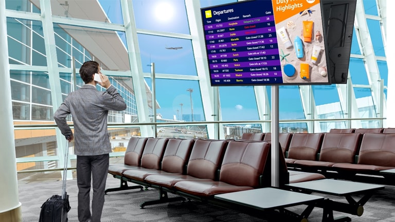 A traveler stands in an airport lounge, checking his phone while looking at a digital departure board displaying flight schedules. The scene highlights the modern, efficient use of signage for real-time updates.