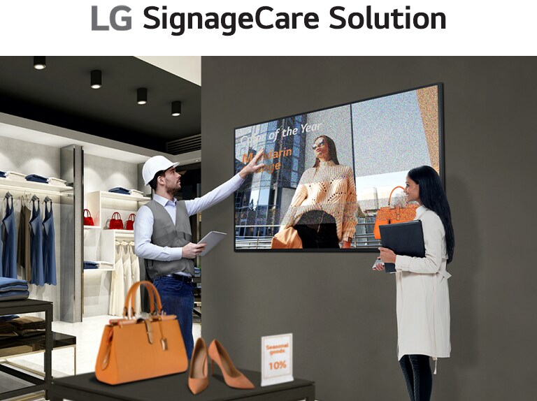 A retail store employee in a vest and hard hat gestures toward a digital display showing a woman with "Employee of the Year" text. Another person, holding a folder, observes the screen in a modern retail space with shelves of clothing and bags.