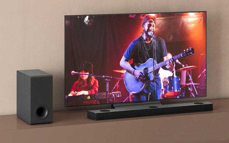 LG TV shows a concert, and LG Sound Bar is placed below LG TV. On the left, sub-woofer is on the brown shelf.