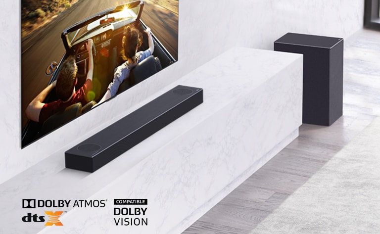 TV is on the wall, LG Soundbar is below on a white marble shelf with a sub-woofer to the right. TV shows a couple in a car. 