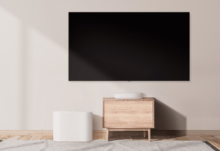 There is a bulky soundbar placed on a wide cabinet and a subwoofer placed on the left. The scene changes and a compact soundbar appears on a smaller cabinet and a subwoofer placed on the left. 