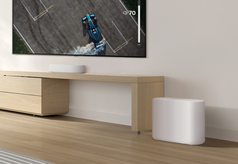 A soundbar is placed on a wooden table, a subwoofer is placed on a wooden floor. A mild sound wave effect comes out from the subwoofer on a floor. There is a TV placed above a soundbar. A racing car on a TV screen is driving very fast and violently and the GUI of TV volume is going up. 