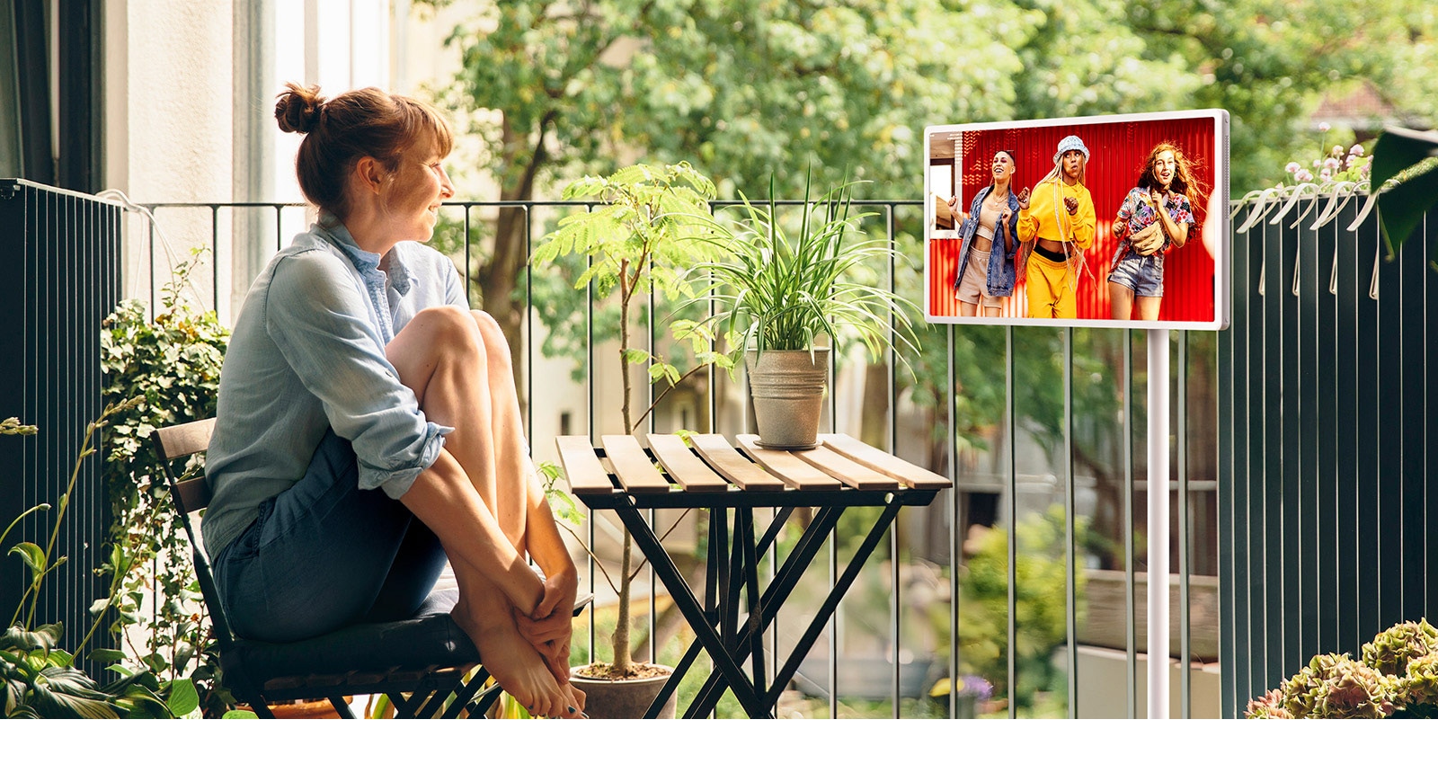 A woman sitting in her outside balcony, watching content on an LG StanbyME 2 on the rollable, adjustable stand.