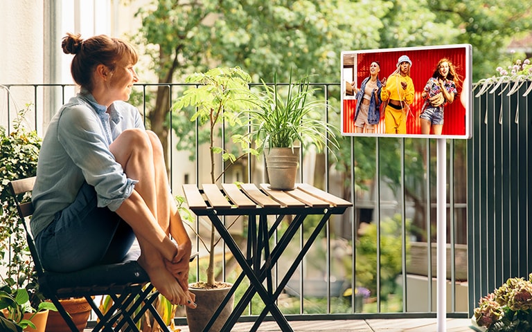 A woman sitting in her outside balcony, watching content on an LG StanbyME 2 on the rollable, adjustable stand.