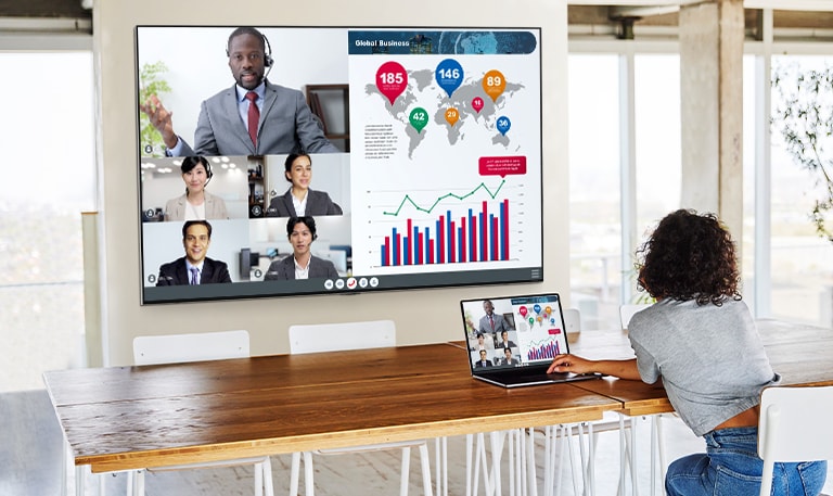 A woman is telecommuting with a laptop and a big tv hung on the wall in the living room.