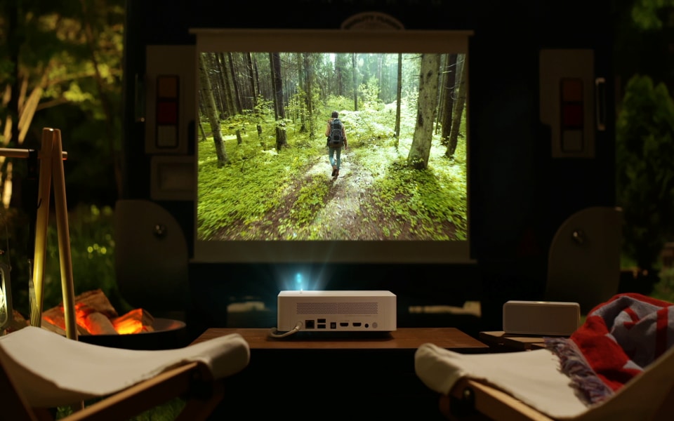 An LG projector showing a forest scene on an outdoor screen with campfire and chairs nearby.