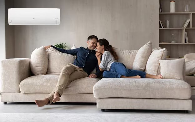 Happy couple relaxing on a beige sofa in a modern living room under an lg air conditioner, smiling at each other.