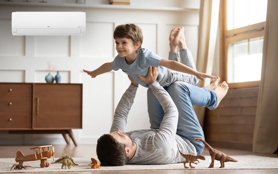 Father playing  with his son on the floor under an LG air conditioner, surrounded by toy dinosaurs and a wooden plane.