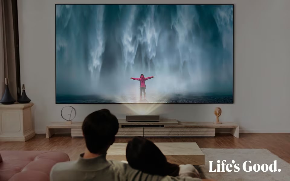 A couple in a living room watching an LG projector display showing a person standing in front of a waterfall.