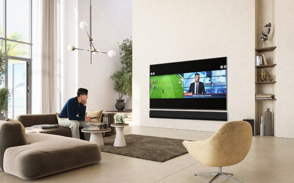 A man sits in a modern, sunlit living room watching a split-screen broadcast on a large wall-mounted LG TV.