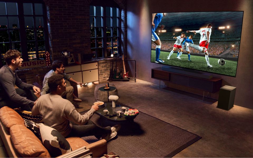 Three men sit on a leather sofa in a dark loft, watching a soccer match on a large widescreen LG TV.