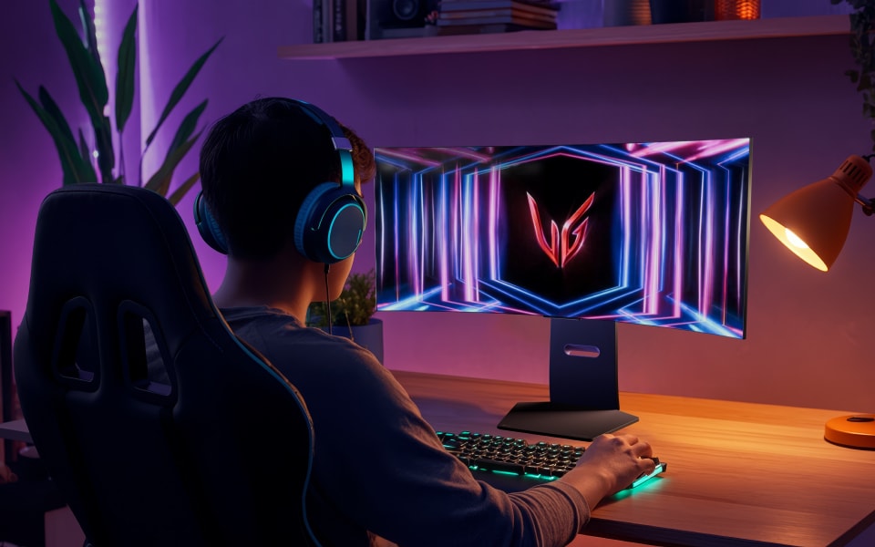 Gamer with headset using RGB keyboard at LG monitor in moody, purple-lit room.