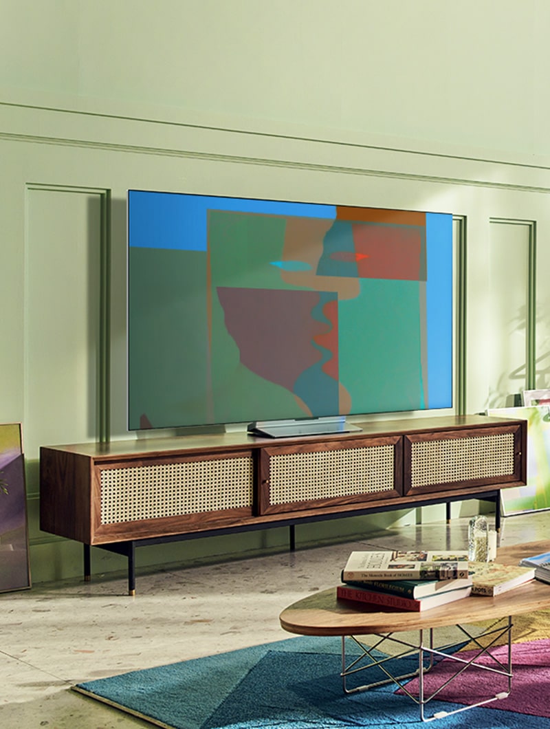 A swivel stand LG OLED television is in a colorful living room with wooden furnishing and greenery. A swivel stand LG OLED television is in a gray and abstract room.