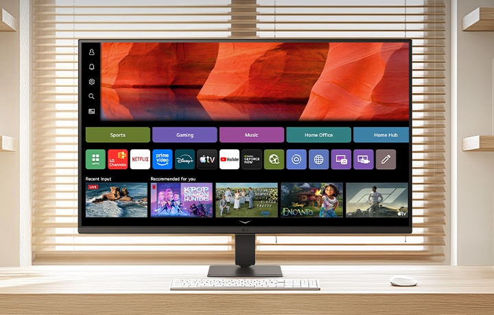 شاشة LG Smart Monitor موضوعة على مكتب خشبي أنيق في مكتب منزلي مشرق، تعرض شاشة webOS الرئيسية مع تطبيقات البث ومربعات المحتوى، إلى جانب لوحة مفاتيح وماوس، ورف كتب وستائر نافذة في الخلفية، مما يسلط الضوء على الاستخدامات الترفيهية والإنتاجية الذكية.