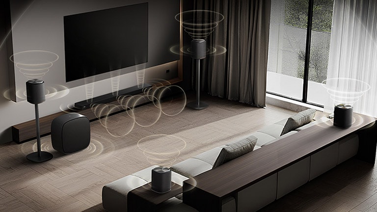 تلفزيون LG TV مع Sound Suite مثبت على الحائط في غرفة معيشة عصرية، مع ترتيب سماعات متعددة حول أريكة حيث تشع الموجات الصوتية المرئية للخارج، مما يوضح إعداد صوت مرن مصمم خصيصًا للمساحة.