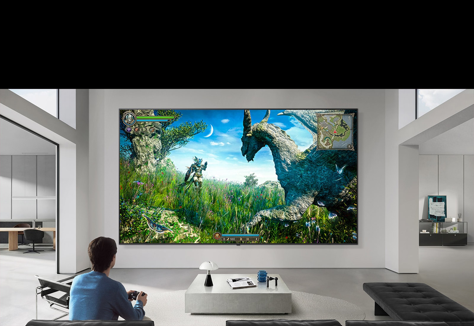 تلفزيون LG TV يظهر مثبتًا على جدار في غرفة معيشة باللونين الأبيض والأسود، ويعرض مشهد لعبة خيالية على شاشة كبيرة بينما يجلس شخص على أريكة تحمل وحدة تحكم.