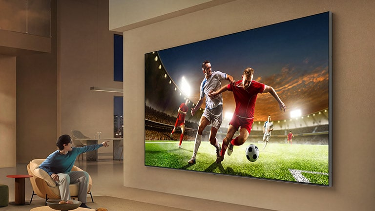 تلفزيون LG للمشاهدة الرياضية مثبت على الحائط في غرفة معيشة فسيحة، مما يعرض مباراة كرة قدم مع اللاعبين أثناء الحركة، بينما يشير شخص جالس إلى الشاشة، مما يوضح عمقًا محسنًا وإحساسًا أكثر انغماسًا بالحركة.