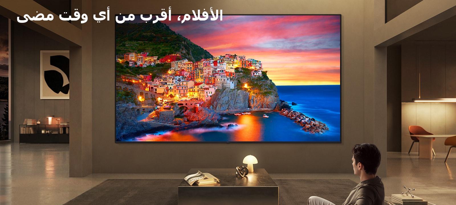 تلفزيون LG Ultra Big TV مثبت على الحائط في غرفة معيشة فسيحة، حيث تعرض الشاشة الواسعة قرية ساحلية نابضة بالحياة عند غروب الشمس، مما يوضح كيف توفر الشاشة الأكبر تجربة مشاهدة سينمائية وغامرة.