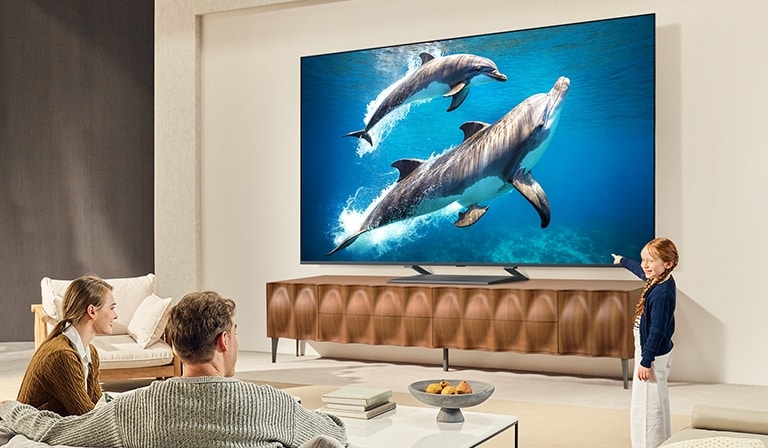 عائلة تجلس على أريكة مواجهة لتلفزيون LG QNED TV مثبت على جدار فوق مكبر الصوت LG Soundbar مع فتاة صغيرة تشير إلى شاشة تصور دلفين. 