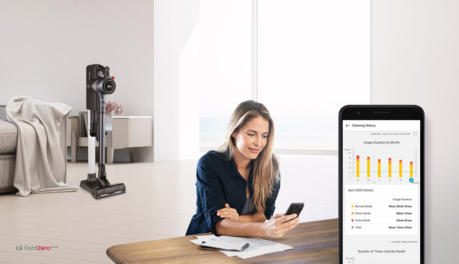 توجد المكنسة الكهربائية في حامل الشحن في غرفة المعيشة في الخلفية مع امرأة تنظر إلى هاتفها في المقدمة. صورة لشاشة الهاتف تُظهر تاريخ تنظيف منتجها.