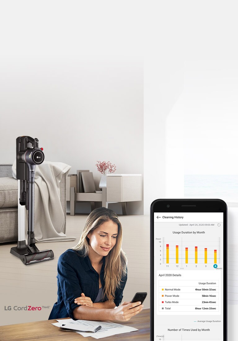 توجد المكنسة الكهربائية في حامل الشحن في غرفة المعيشة في الخلفية مع امرأة تنظر إلى هاتفها في المقدمة. صورة لشاشة الهاتف تُظهر تاريخ تنظيف منتجها.
