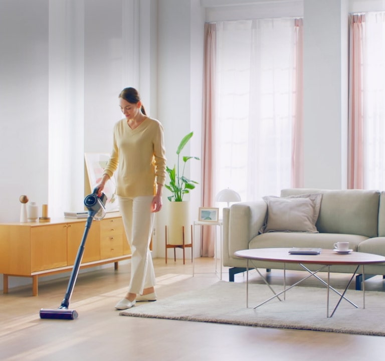 امرأة تقف في غرفة معيشة ذات أرضية صلبة وسجادة وتنظف الأرضية بالمكنسة الكهربائية.