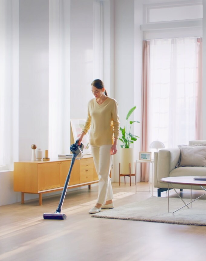 امرأة تقف في غرفة معيشة ذات أرضية صلبة وسجادة وتنظف الأرضية بالمكنسة الكهربائية.