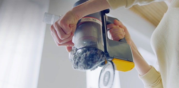 تقبض المرأة على مقبض المكنسة الكهربائية وتستخدم اليد الأخرى لتحريك الرافعة لإفراغ العلبة بسهولة.