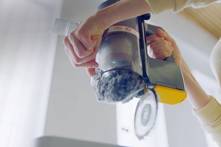 تقبض المرأة على مقبض المكنسة الكهربائية وتستخدم اليد الأخرى لتحريك الرافعة لإفراغ العلبة بسهولة.