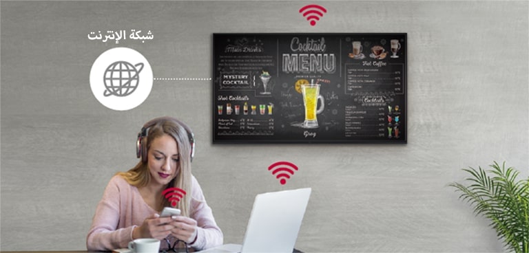 سلسلة UR3J على الحائط وامرأة تستخدم كمبيوتر شخصي وهاتف جوال. تظهر هذه الصورة أن اللافتة يمكن توصيلها لاسلكيًا بجهاز كمبيوتر شخصي والهاتف الجوال الذي تحمله المرأة.