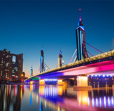 صورة ليلية لجسر حديث وناطحات سحاب ذات إضاءة ساطعة تنعكس في الماء، مما يوضح التباين العالي وتفاصيل اللون.