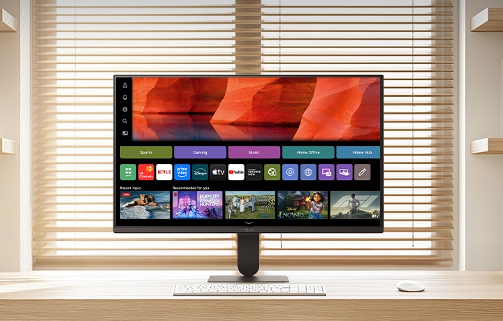 شاشة LG Smart Monitor موضوعة على مكتب خشبي أنيق في مكتب منزلي مشرق، تعرض شاشة webOS الرئيسية مع تطبيقات البث ومربعات المحتوى، إلى جانب لوحة مفاتيح وماوس، ورف كتب وستائر نافذة في الخلفية، مما يسلط الضوء على الاستخدامات الترفيهية والإنتاجية الذكية.