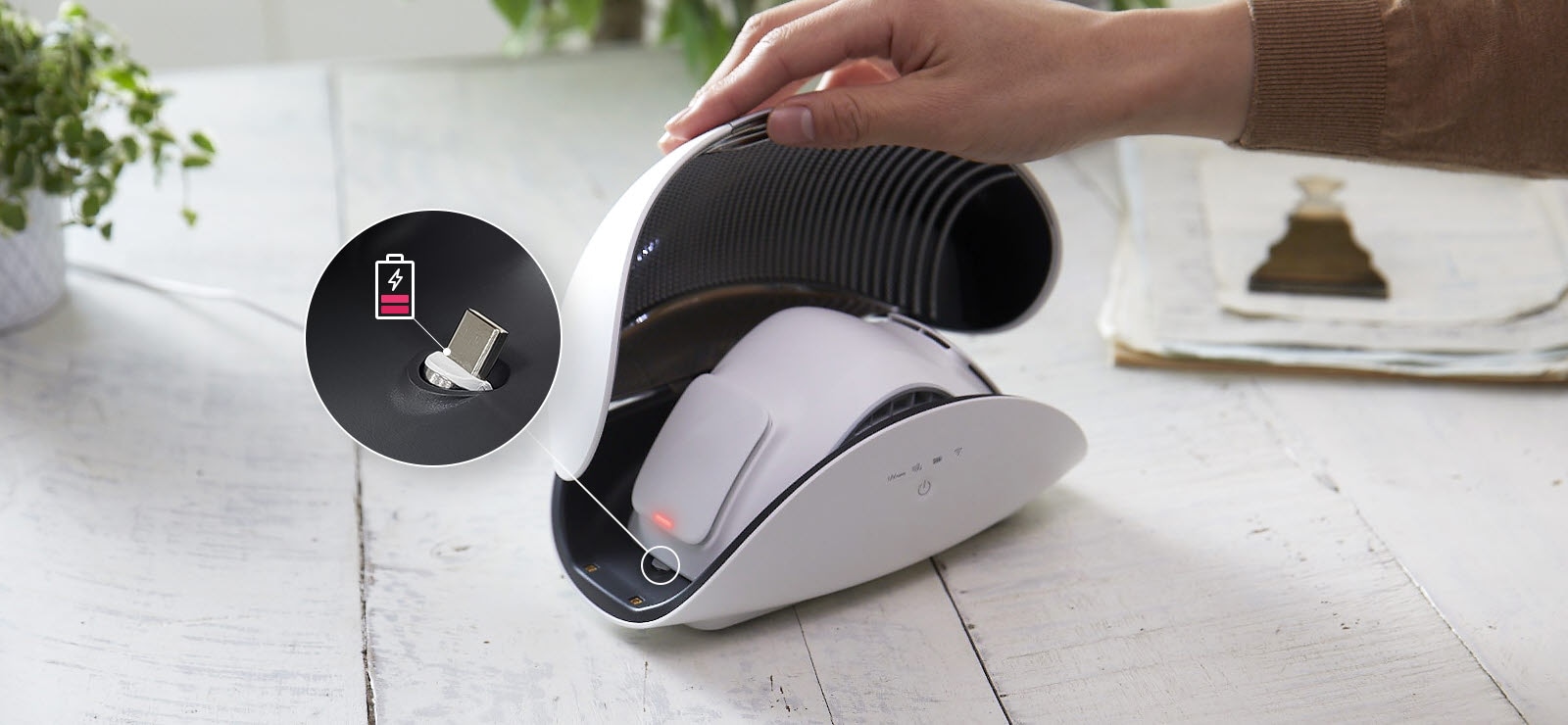 يد تمسك بغطاء علبة جهاز تنقية لإظهار جهاز تنقية الهواء القابل للارتداء LG PuriCare الموجود بالداخل.  يوجد ضوء أحمر على جانب جهاز تنقية الهواء القابل للارتداء بالداخل، وتظهر الصورة في دائرة بها خط على جانب القناع منظرًا مكبّرًا للمكان الخاص بإعادة شحن القناع.  يوجد أيضًا رمز بطارية أحمر اللون وشبه فارغ يشير إلى أن الوقت قد حان لتوصيله. 