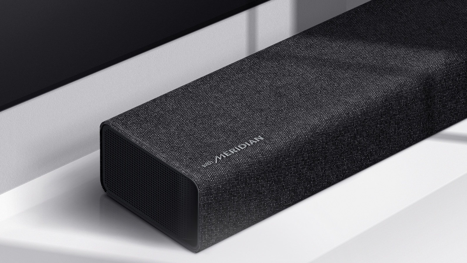 لقطة مقرّبة للجانب الأيسر من مكبرات الصوت LG Soundbar مع شعار Meridian الظاهر في الزاوية اليسرى السفلية من المنتج. يظهر الجانب الأيسر السفلي من التلفزيون أيضاً.