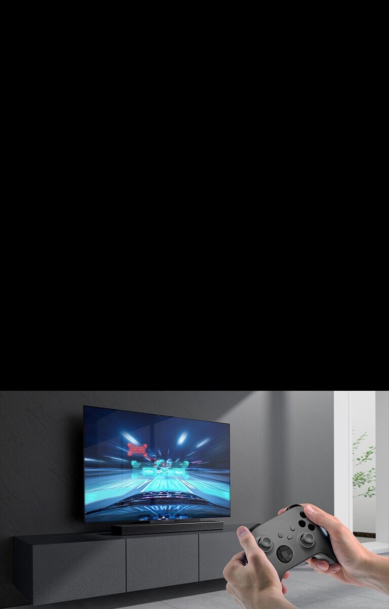 مكبر الصوت على الخزانة ويظهر مشهد لعبة السباق على التلفاز المتصل بمكبر الصوت. وحدة تحكم في الألعاب في الجانب الأيمن السفلي من الصورة وتمسك بها يدان.