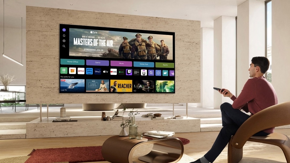 A man uses a Magic Remote with a large LG smart TV displaying a home screen with various streaming apps.