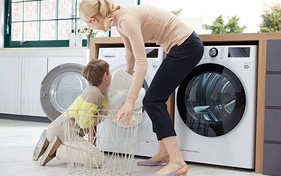A woman and child are doing laundry, loading clothes into a white LG washing machine beside a matching dryer.