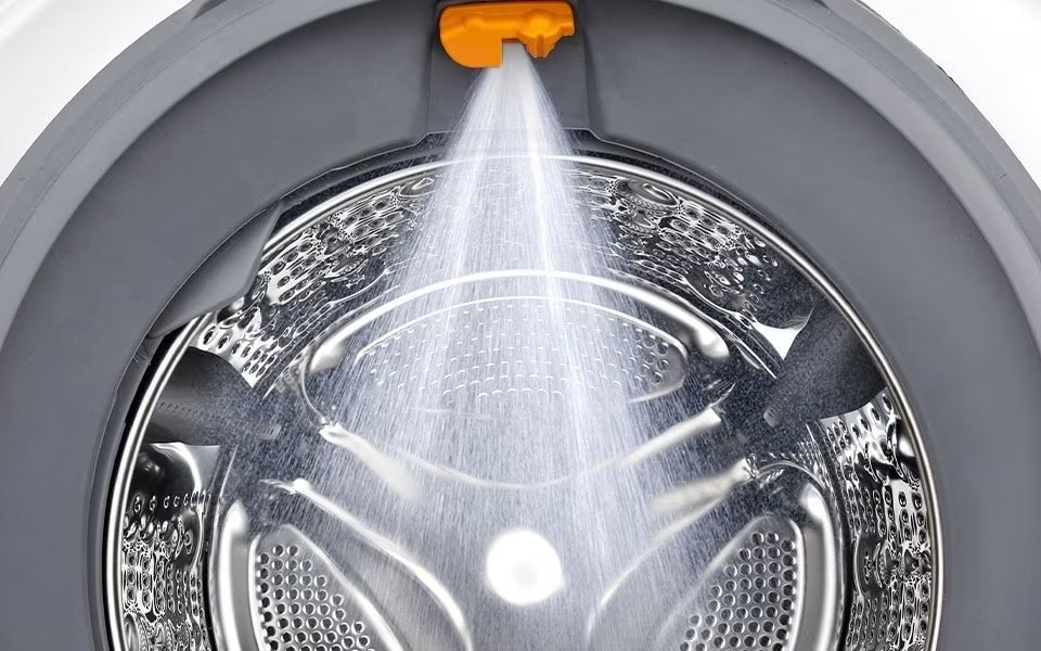 A close-up inside an LG washing machine with water spraying from an orange nozzle onto the stainless steel drum.