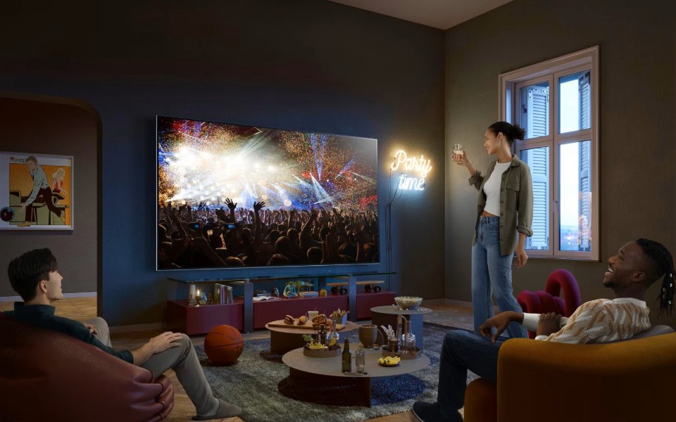 ثلاثة أصدقاء يشاهدون حفلاً موسيقياً على تلفزيون إل جي في حفلة مع وجبات خفيفة ولافتة نيون مكتوب عليها 