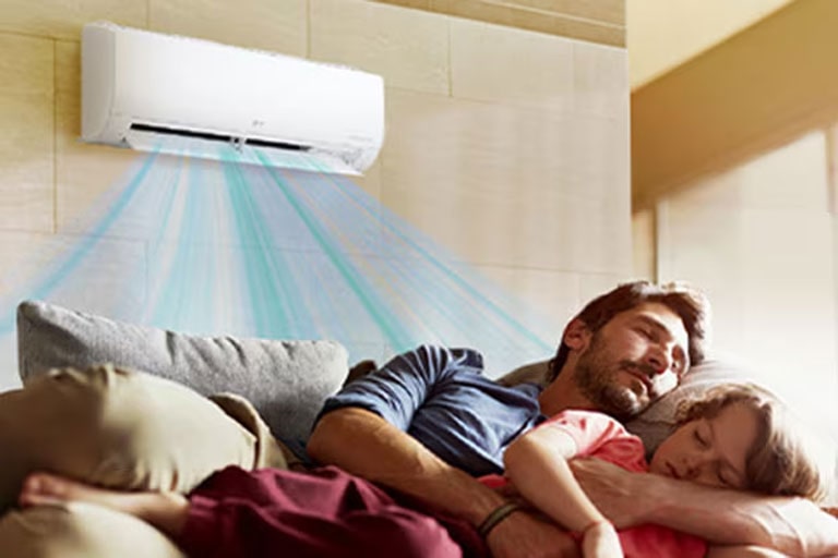 A father and daughter sleep on a couch beneath an air conditioner that is blowing air out over them.