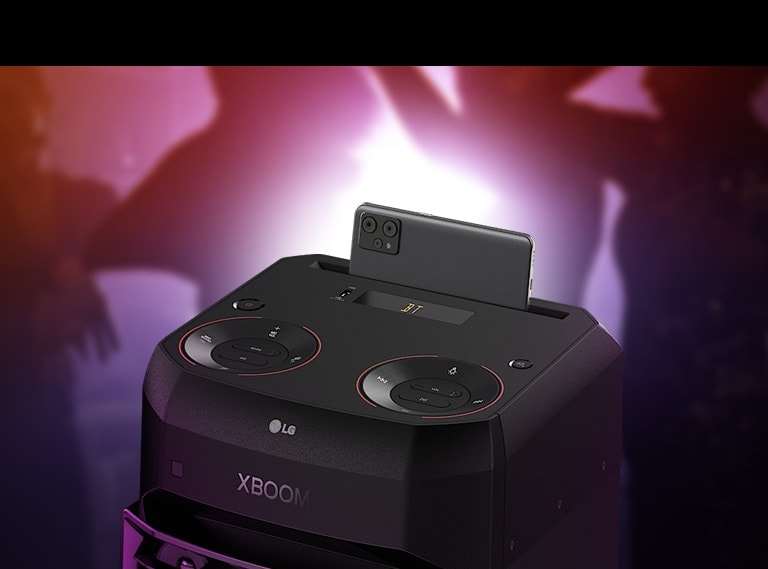 LG XL2S A close-up view of the top of LG XL2S, highlighting on the phone cradle. A smartphone is placed on the phone cradle. Behind, there are silhouettes of people dancing.