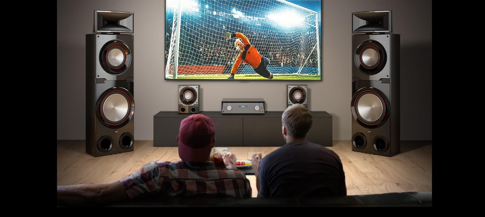 LG MR11 is placed in the living room. LG TV is hung on the wall, between two giant tall boy speakers, displaying a soccer game. Two people are watching the game.