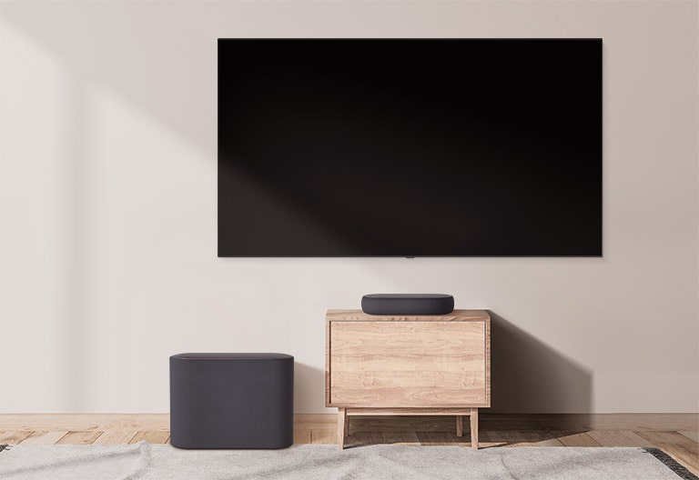 There is a bulky soundbar placed on a wide cabinet and a subwoofer placed on the left. The scene changes and a compact soundbar appears on a smaller cabinet and a subwoofer placed on the left. 