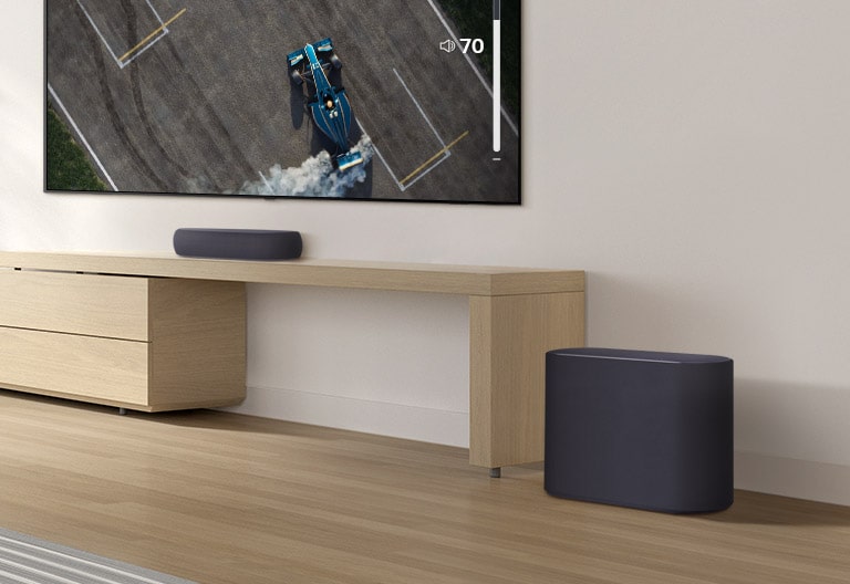 A soundbar is placed on a wooden table, a subwoofer is placed on a wooden floor. A mild sound wave effect comes out from the subwoofer on a floor. There is a TV placed above a soundbar. A racing car on a TV screen is driving very fast and violently and the GUI of TV volume is going up. 