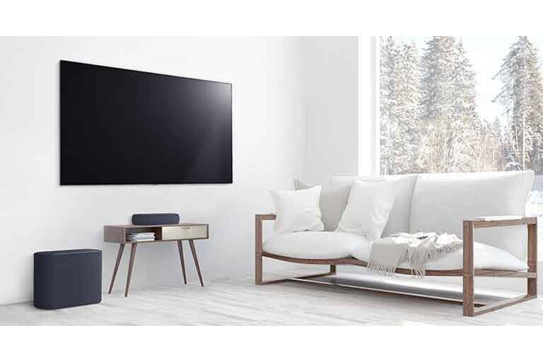 A soundbar is placed on a small tea table,  a subwoofer placed on the left side and TV is placed above a soundbar. The room is overall white. 