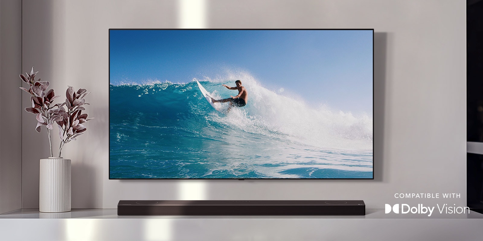TV is on the wall. TV shows a man surfing on big wave. LG Soundbar is right below TV on a white shelf. There is a vase with a flower right next to the soundbar. (play the video) 